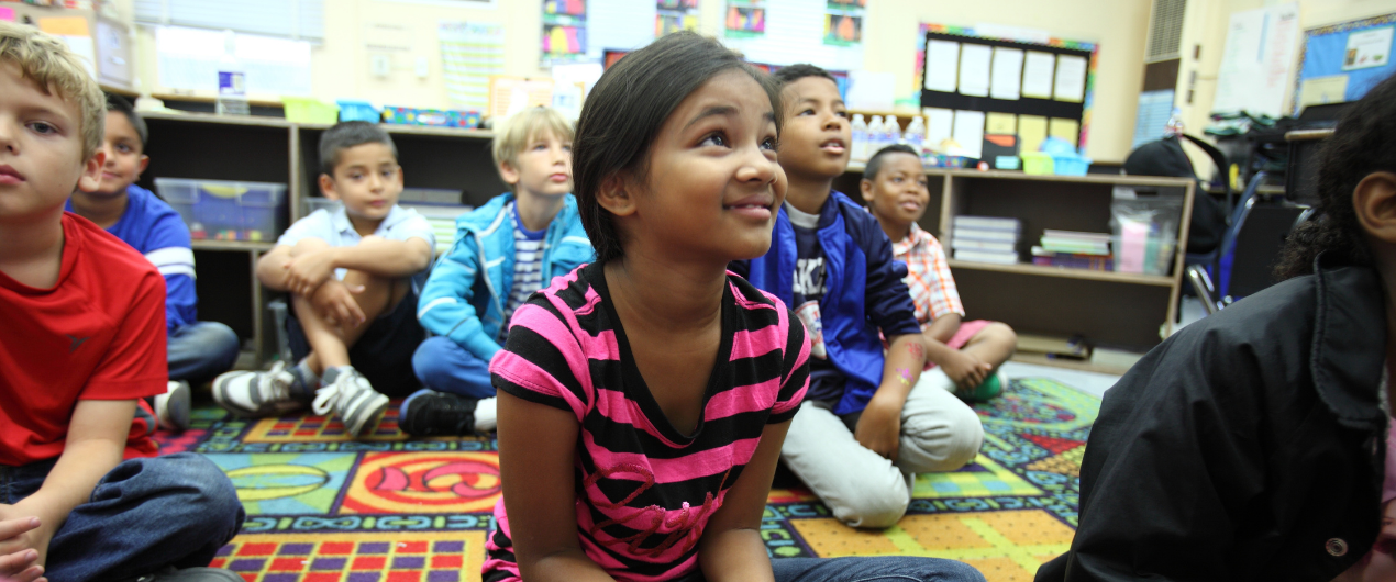 Picture of young girl sitting on carpet in her classroom with other boys and girls sitting near her
